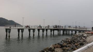 O deck do pescador visto de longe, com enrocamento na praia em primeiro plano. #Paratodosverem