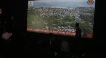 homem está à frente de telão onde está projeta imagem de morr. No pé da imagem se lê Como construir um morro mais seguro. #paratodosverem