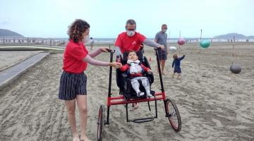 Criança com deficiência anda em bicicleta acessível, guiada por adultos, na faixa de areia. #paratodosverem