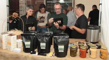 estande de café em primeiro plano, com pacotes do grão moído, e pessoas atrás, bebendo café. #paratodosverem 