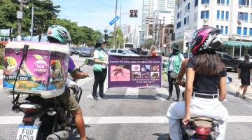agentes seguram faixa com informações sobre o mosquito da dengue sob semáforo fechado de avenida. Motos estão paradas em primeiro plano. #paratodosverem 