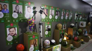 Bolas de futebol estilizadas estão expostas em museu sobre pedestais. Atrás, em painel, estão fotos dos designers de cada peça. #Paratodosverem