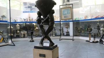 troféu no centro de uma sala com vários troféus espalhados. Peça tem dois homens segurando um troféu ao mesmo tempo. #paratodosverem