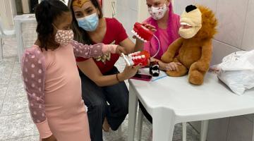 criança usando máscara mexe em bicho de pelúcia que está sobre uma mesa. O ambiente está decorado com balões de gás cor-de-rosa. Ao lado da menina há duas mulheres, uma deles usa uma fantasia infantil. #paratodosverem