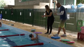 Mulher está dentro da piscina e conversa em Libras com tradutora que está do lado de fora ao lado do professor de natação. Pracegover