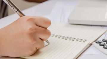 close de mão segurando caneta e escrevendo em caderno. Há uma calculadora e um notebook sobre a mesa. #paratodosverem