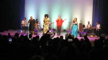 plateia lotada assiste mulheres cantando no palco. #paratodosverem