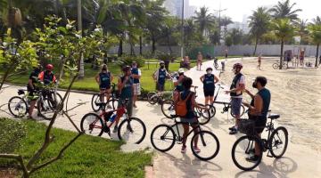 Grupo de ciclistas está reunido, parado, no jardim da orla. #Paratodosverem