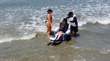 jovem está sentada em cadeira anfíbia tomando banho de mar. Técnico controla a cadeira. A mãe da jovem está em pé ao lado. Eles estão no mar. #paratodosverem 
