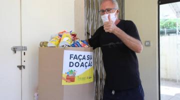 Homem usando máscara deposita alimento em caixa de papelão. Na frente da caixa está escrito 