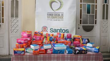 mesa com diversos pacotes de absorventes. Banner do fundo social ao fundo. #paratodosverem