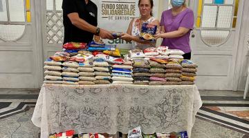 um homem e duas mulheres estão atrás de uma mesa cheia de pacotes de alimentos. No chão também há duas caixas cheias de alimentos. Ao fundo, um banner do fundo social de solidariedade. #paratodosverem