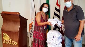 mulher e homem estão em pé, lado a lado, ambos segurando camisetas. Há uma pilha de camisetas sobre uma cadeira. Ao lado deles há uma tribuna onde estão as letras acdbs. #paratodosverem