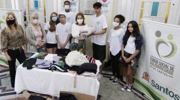 mesa cheia de roupas e cobertores com estudantes e adultos atrás posando para a foto. Há um banner do fundo social de solidariedade 