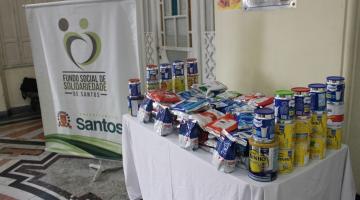 pacotes e latas de leite em pó sobre uma mesa. Ao lado, um banner do fundo social de solidariedade. #paratodosverem