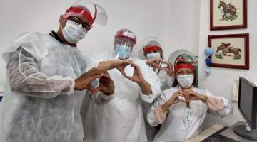 Quatro funcionários do setor de saúde estão usando avental, mascaras cirúrgicas e de acetato e gorro. Eles fazem gesto de coração com as mãos posando para a foto. #Paratodosverem