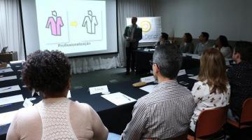 Profissionais estão reunidos em sala numa mesa em forma de U. Ao fundo um homem fala ao grupo. Há um telão que projeto a imagem de dois corpos desenhados. #Pracegover