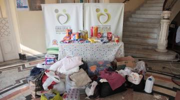 produtos de higiene e roupas estão sobre uma mesa e empacotados em torno dela. Atrás da mesa há um banner do fundo Social de solidariedade. #paratodosverem