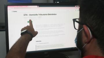 homem está de costas para foto apontando dedo para tela de desktop aberto n a página do Domicílio eletrônico. #paratodosverem