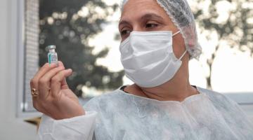 mulher segura frasco com dose de vacina. Ela usa avenal, touca e máscara. #paratodosverem
