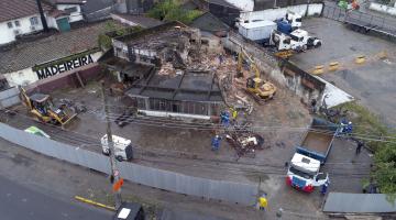 Imagem aérea de antigo posto em demolição. Há máquinas e caminhões na área. Parte da estrutura, ao fundo, já está derrubada. #Pracegover