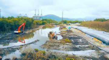 vista geral de canal máquinas para desassoreamento. Do lado esquerdo, vegetação. Ao fundo, guindastes do porto. #paratodosverem