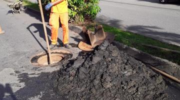 operário uniformizado segura uma longa ferramenta que está dentro de um poço de visita em Santos. Ao lado, na pista há um acumulado de lama. #paratodosverem