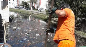 homem limpa trecho de canal, do lado de fora, com auxílio de uma ferramenta de cabo longo