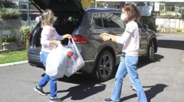 Mulher entrega sacolas com doações para voluntária, com carro ao fundo. 