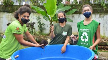 Três jovens seguram terra e local de compostagem #paratodosverem