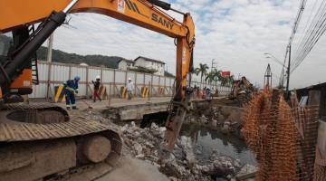 Máquinas e homens trabalham na construção de pontilhão. A máquina está removendo parte do piso. Ao redor se vê um trecho de rio. #Pracegover