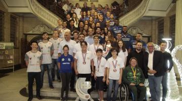 Atletas, técnicos e autoridades municipais posam para foto na escada do hall da Prefeitura, atrás do troféu. #Pracegover