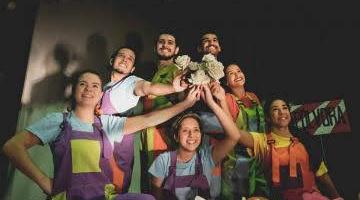 Alunos da escola de artes cênicas posam para foto com figurinos coloridos. Há quatro pessoas em pé e três sentadas. todos estão com as mãos estendidas que se juntam em um centro. #Pracegover