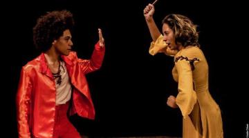 dois artistas estão no palco frente a frente. O homem, à esquerda, faz um gesto de pare para mulher, que segura uma colher de pau como se fosse agredi-lo. #paratodosverem