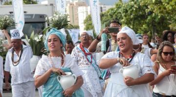 Mulheres vestidas com roupas típicas de umbanda, com turbantes e vasos nas mãos caminham pela praia. #Pracegover