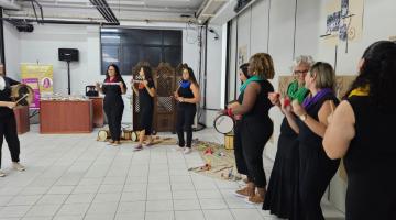 mulheres estão em semicírculo em pé tocando instrumentos de percussão. #paratodosverem