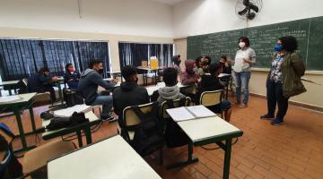 sala de aula com alunos sentados e duas mulheres em pé. Uma delas fala ao grupo. #paratodosverem