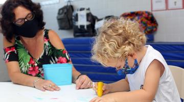 Educadora está ao lado de criança que desenha sobre uma mesa. #paratodosverem