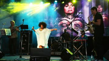 cantora está no palco, se movimentando. Banda segue. Ao fundo, no telão, uma imagem de Elza Soares. #paratodosverem