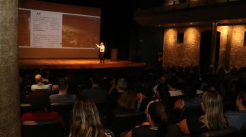 Servidores sentados na plateia do teatro assistem exibição de secretário, em pé no palco, abaixo de telão. #pratodosverem