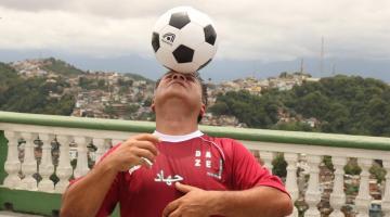 homem está equilibrando bola na cabeça.Ele está à frente de uma mureta no alto de um morro. Ao fundo, se vê as casas e a vegetação em outro morro. #paratodosverem