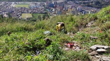 Homem retira lixo de área do morro com muita mata e cidade ao fundo. #paratodosverem