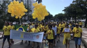 pessoas caminhando na avenida, usando roupas amarelas. Algumas seguram balões amarelos em alusão à endometriose. Duas pessoas à frente da marcha seguram uma faixa onde se lê Endomarcha. #paratodosverem