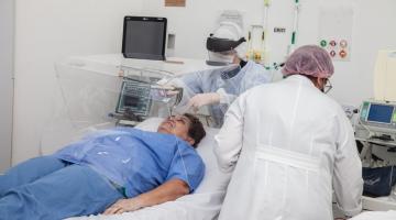 Dois profissionais de saúde uniformizados estão atendendo um paciente no leito de hospital. #paratodosverem