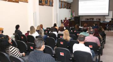 Pessoas sentadas em um auditório observam pessoa falando e telão ao fundo #paratodosverem