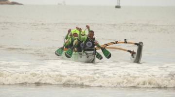 equipe em canoa havaiana vem chegando na beira da praia. Ao fundo, apenas o farol no meio do mar. #paratodosverem