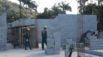 Operários assentam blocos de parede em canteiro de obras. Há estruturas de ferro implantadas no solo. As paredes têm cerca de dois metros de altura. #Paratodosverem