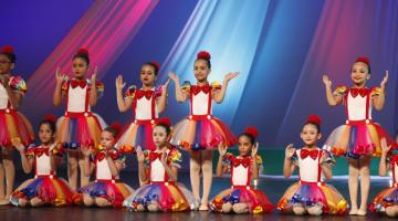 meninas estão usando a mesma roupa em apresentação de balé. Algumas estão sentadas e outras em pé no palco. #paratodosverem