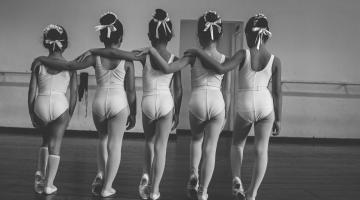 Meninas estão vestidas para apresentação de bale. Todas estão de costas para a foto, cada uma com o braço esquerdo no ombro da outra; os pés estão em movimento de balé. #Pracegover