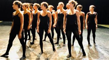 #pracegover Quinze bailarinas vestidas de negro posem de pé olhando para o lado direito 